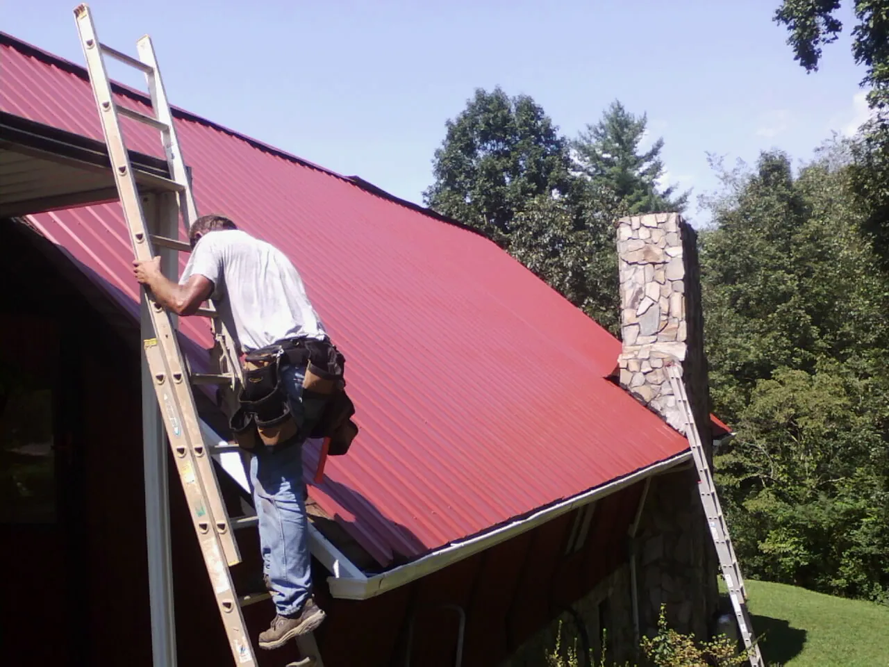 Roofer on a ladder beside a red metal roof and stone chimney — Soffit & Fascia Repair by Roofing Brothers in Schuyler